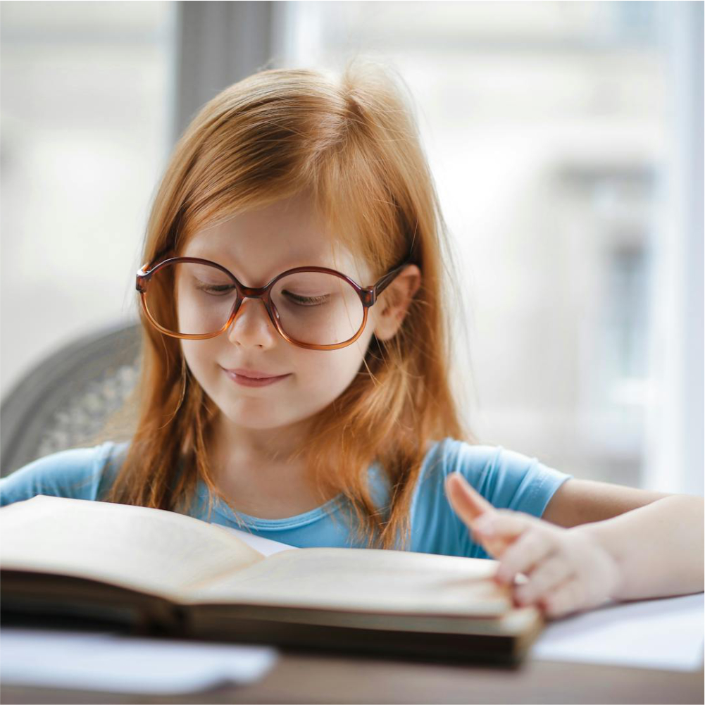 Niña pequeña con gafas leyendo un libro durante una valoración visual infantil.